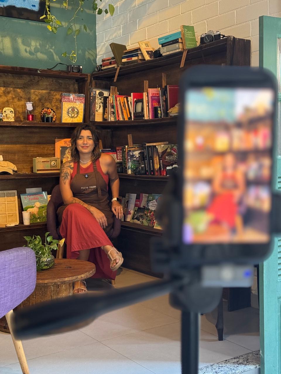 Gabi Machado, de avental da Feito Trama, sentada diante de uma estante de livros